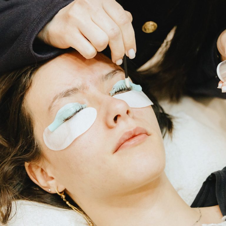 Portrait of a young unrecognizable Caucasian girl cosmetologist in a dark jacket who washes off brown paint from an eyelash on a silicone roller of the right eye using a microbrush to a teenage client lying on a cosmetology table in a home beauty salon for eyelash lifting, top side close-up view with selective focus.