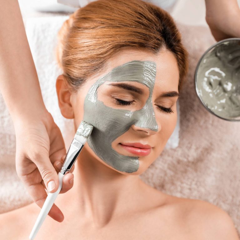Cosmetologist applying mask onto woman's face in spa salon, top view